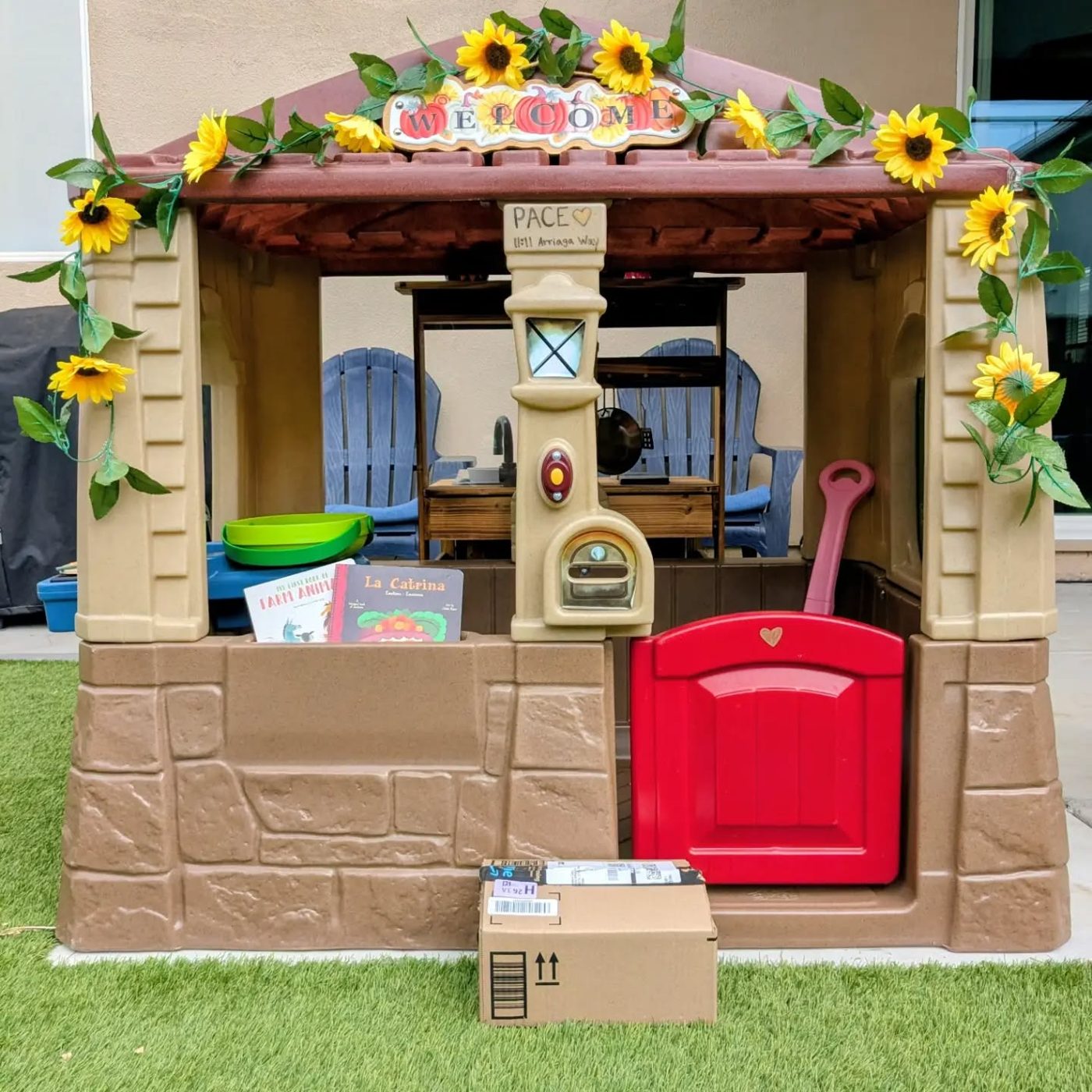 A backyard playhouse trimmed with sunflowers and a welcome sign, with picture books in the window